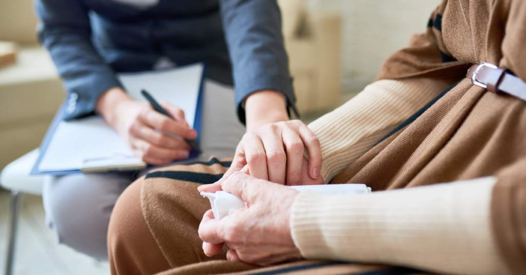 rimer plano de dos personas, una de las cuales parece ser un profesional, posiblemente un terapeuta o consejero, tomando notas en un portapapeles. La otra persona sostiene un pañuelo y tiene una mano colocada sobre la suya, sugiriendo un gesto de apoyo o consuelo. El profesional lleva un blazer oscuro y pantalones de color claro, mientras que la otra persona viste un conjunto marrón y beige con un cinturón. La escena sugiere un entorno de apoyo emocional en terapia