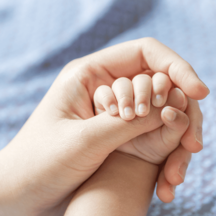 Primer plano de la mano de una madre sosteniendo la mano de un bebé. El adulto está sujetando delicadamente la mano del bebé, y los dedos del bebé están envueltos alrededor de uno de los dedos del adulto. El fondo está desenfocado y parece ser una tela suave de color azul claro. Esta imagen simboliza cuidado, protección y el vínculo entre la madre su bebé.